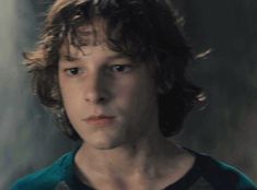 a young man with curly hair and blue eyes stares into the camera while standing in front of a dark background