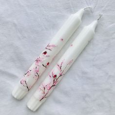 two white candles with pink flowers painted on them sitting on a bed sheet next to each other