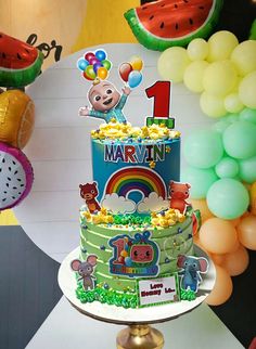 a birthday cake that is on top of a table in front of balloons and watermelon