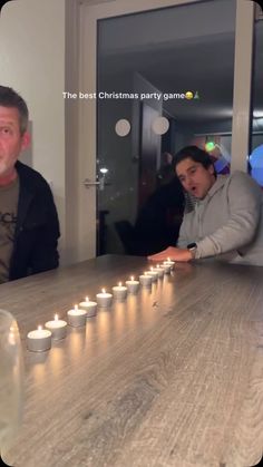 two people sitting at a table with candles in front of them and one person standing next to the table