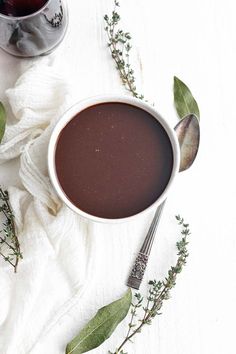 a cup of hot chocolate next to two silver spoons and some green leaves on a white cloth