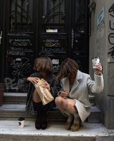 two women sitting on the steps taking pictures with their cell phones in front of graffiti covered doors