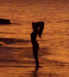 a woman standing in the water at sunset with her back to the camera and arms behind her head