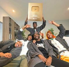 four people sitting on a bed with their arms in the air and one person holding up a cardboard box