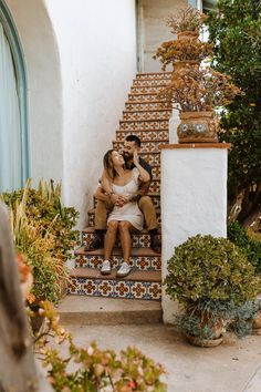 a man and woman are sitting on the steps in front of a house with potted plants