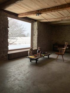 two children are sitting on benches in an empty room with large windows and stone walls