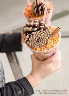 two people holding up cups filled with food and pine cones on top of each cup