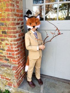 a man in a suit and fox mask standing next to a brick wall holding a twig