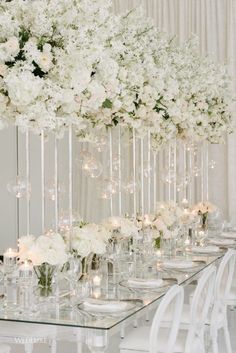 a long table with white flowers and candles in vases on the top is surrounded by clear glassware