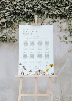 an easel with a seating chart on it in front of a plant covered wall