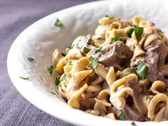 a white bowl filled with pasta and meat on top of a purple table cloth next to a fork