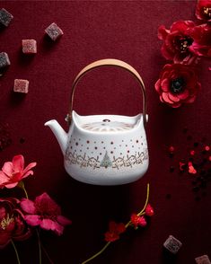 a white tea pot sitting on top of a table next to red flowers and sugar cubes