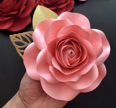 a hand holding a paper flower in front of red and pink flowers on a black table