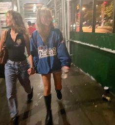 two women walking down the sidewalk in front of a building at night, one is wearing an old school style jacket and the other wears high heeled boots