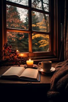 an open book, coffee cup and candle sit on a table in front of a window