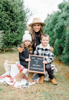 a woman and two children sitting on a blanket in front of christmas trees holding a sign that says, wish you merry christmas