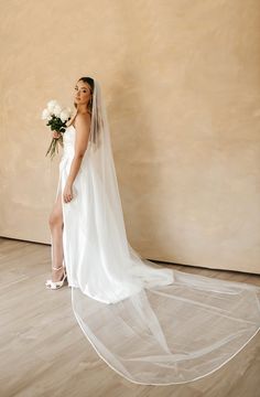 a woman standing in front of a wall wearing a wedding dress and holding a bouquet