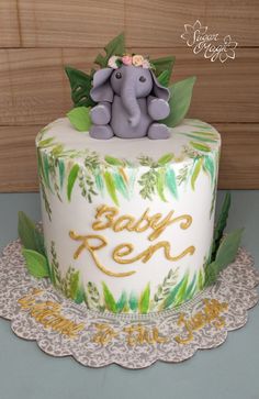 a baby shower cake with an elephant on top and leaves around the edges is decorated with gold lettering