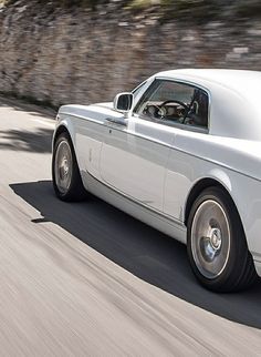 a white rolls royce driving down the road in front of a rock wall and trees