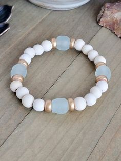 This stacker boasts beautiful white wooden and gold beads, alongside environmentally friendly recycled glass from the Krobo tribe. Embrace a boho and neutral aesthetic while making a positive impact with this stylish piece.    Cream colored wooden beads   Gold heishi colored beads   Aqua Recycled glass   Fits a wrist up to 6.5"   Created in studio - Wilmington, NC White Recycled Glass Jewelry For The Beach, White Stretch Bracelet With Round Wooden Beads, White Bracelets With Round Wooden Beads, White Wooden Beads Bracelet, Layered Bracelets Aesthetic, Wood Bead Bracelet, Neutral Aesthetic, Wood Bracelet, Gold Armband