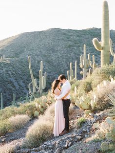 a man and woman standing next to each other in front of a cactus covered hill