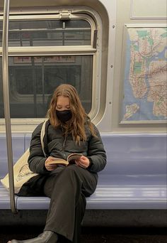 a woman sitting on a subway reading a book and wearing a mask while looking at her phone