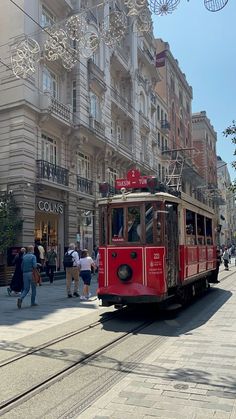 a red trolley car traveling down a street next to tall buildings and people walking on the sidewalk