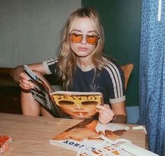 a woman sitting at a table with her book and sunglasses on, reading the magazine