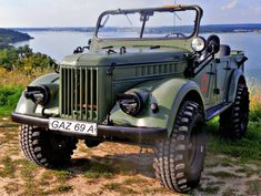 an old green jeep parked on the side of a hill next to a body of water