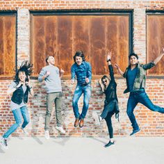 five people jumping in the air near a brick wall with windows and doors behind them