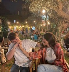 two people sitting on wooden chairs in an outdoor area at night, one holding his face to the other's mouth