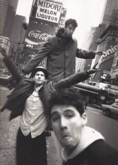 three men standing on the side of a road in front of a neon sign and tall buildings