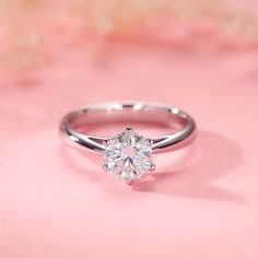 a white gold engagement ring with a round brilliant cut diamond in the center on a pink background