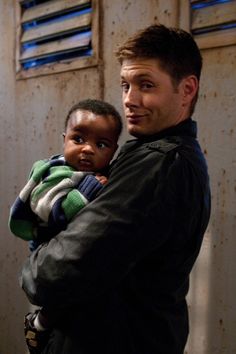 a man holding a small child in his arms while standing next to a wall with shutters