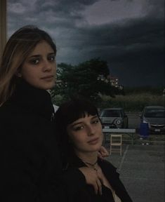 two women standing next to each other in front of a building with dark clouds behind them