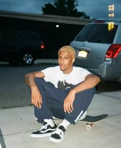 a young man sitting on the sidewalk with his skateboard in front of him and cars behind him