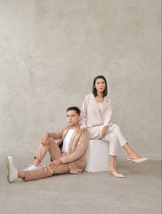 a man and woman sitting next to each other in front of a concrete wall, posing for the camera