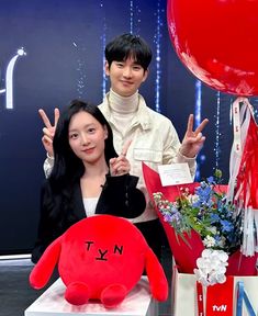 a man and woman standing next to each other in front of a red stuffed animal