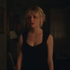 a woman with blonde hair wearing a black dress and looking at the camera while standing in a dark room