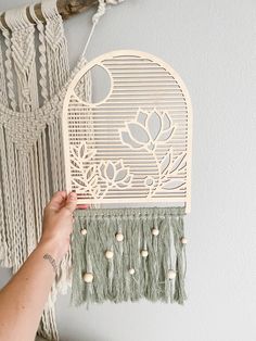 a person is holding up a wall hanging with flowers and leaves on it, next to a macrame