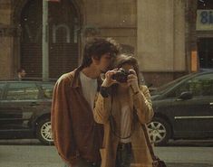 a man and woman standing next to each other on the street taking pictures with their cell phones