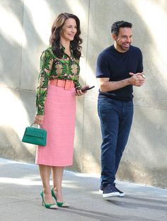 a man standing next to a woman in a pink dress and green purse on the street