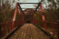 an old rusty red bridge in the middle of autumn with leaves on the ground and trees around it