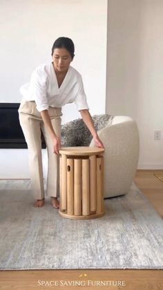 a woman in white shirt and beige pants standing next to a table with a stool on it