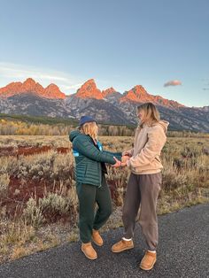 two women standing on the side of a road with mountains in the background