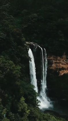 a large waterfall in the middle of a forest with people standing at it's base