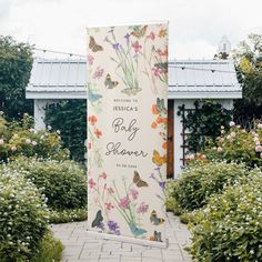 a sign with flowers and butterflies on it in the middle of a walkway surrounded by bushes