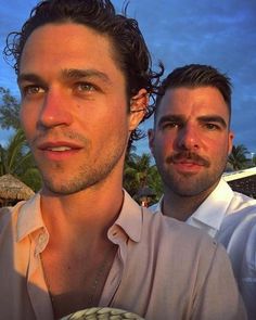 two men standing next to each other in front of some palm trees and blue sky