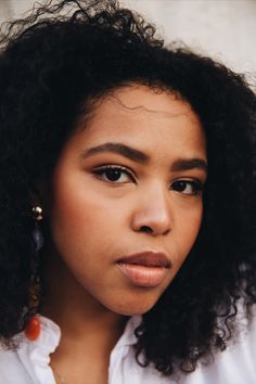 a close up of a person wearing earrings and a white shirt with curly hair, looking at the camera