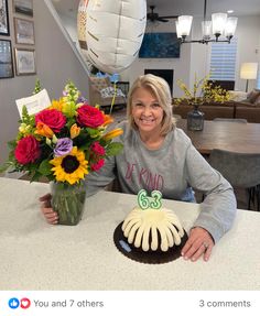 a woman sitting at a table with a cake and flowers in front of her smiling